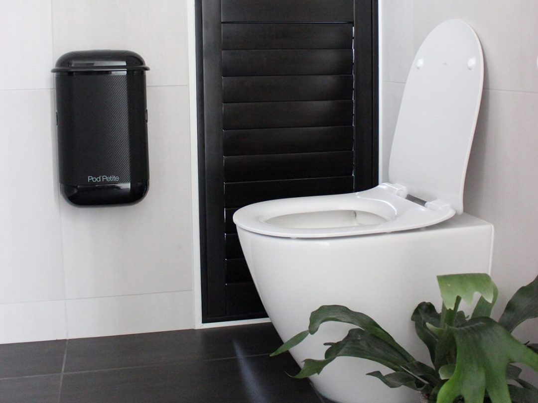 A black Pod Petite sanitary disposal unit beside toilet with a Pod Wrap Carbon Fibre decal.