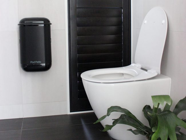 A black Pod Petite sanitary disposal unit beside toilet with a Pod Wrap Carbon Fibre decal.