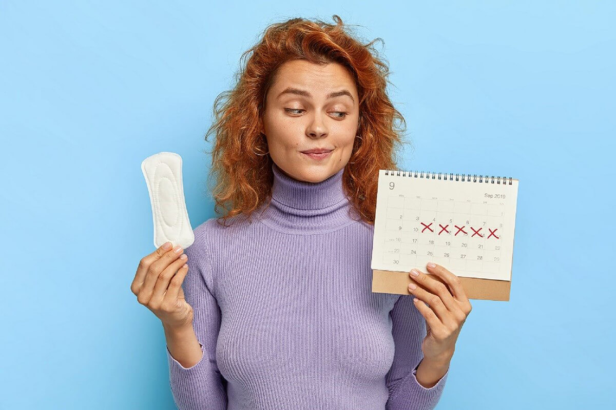 A woman holding a sanitary pad in one hand and a calendar with her period dates crossed off in another.