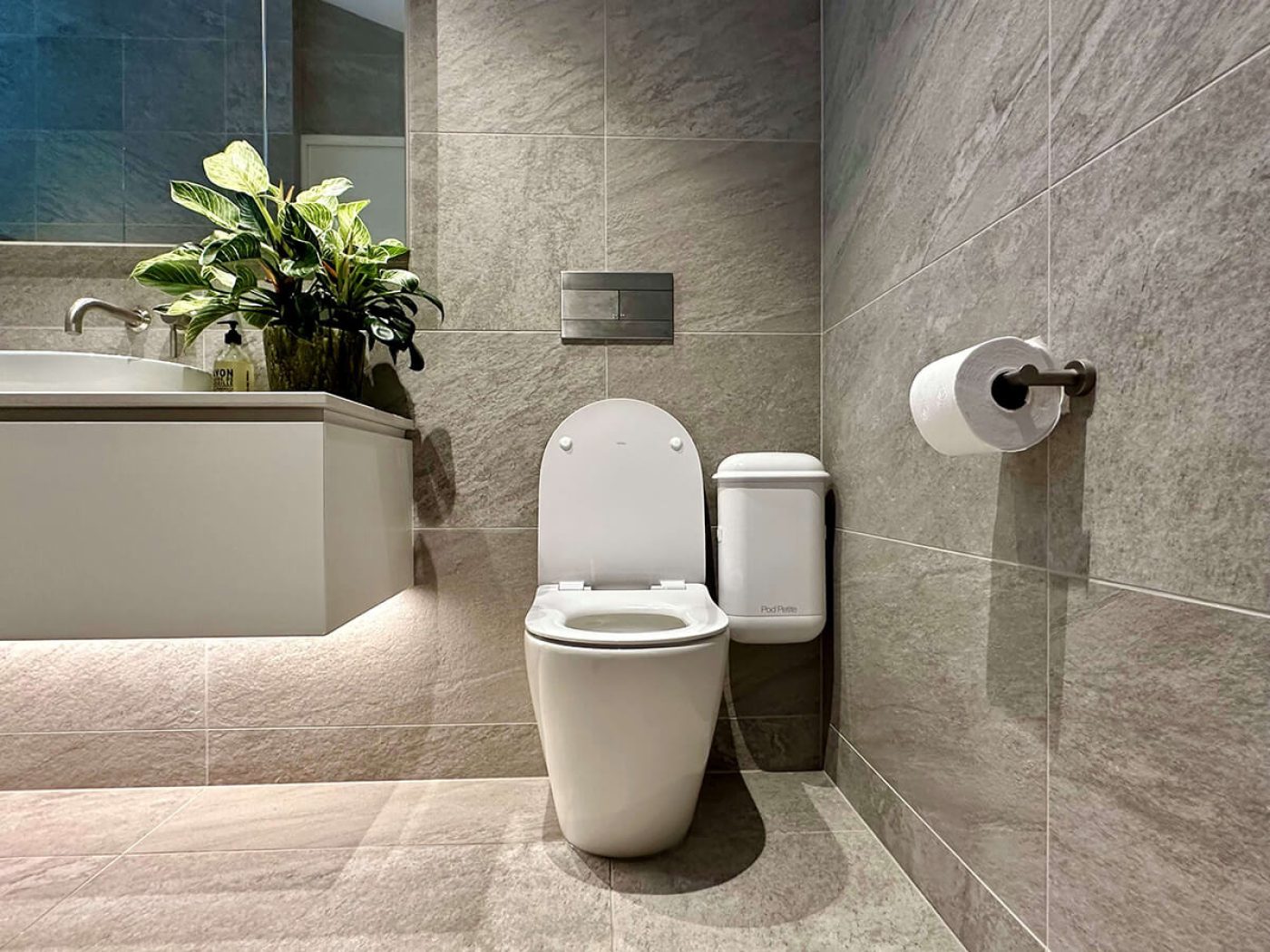 A Pod Petite Auto white sanitary disposal unit in a grey tiled bathroom.
