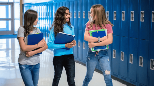 Feminine Hygiene Products in Schools – 3 school girls walking down the hallway, smiling, with blue lockers behind them