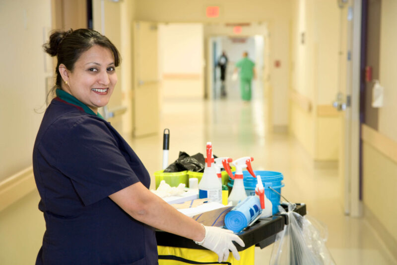 Environmental Services Custodian with a trolley full of cleaning supplies in hospital corridor